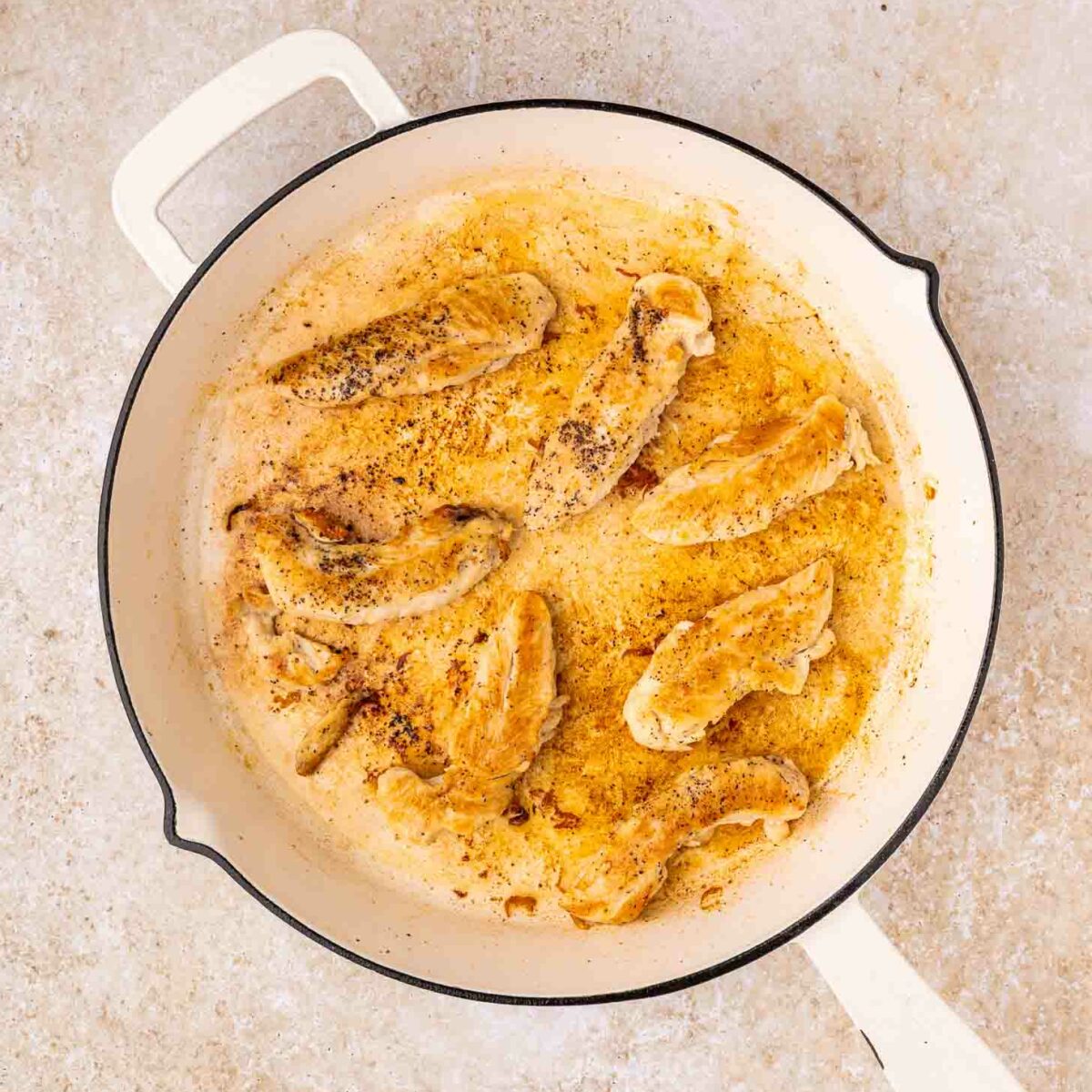 Cooked seasoned chicken strips in a white skillet on a light-colored countertop. The chicken is browned and surrounded by juices and spices.