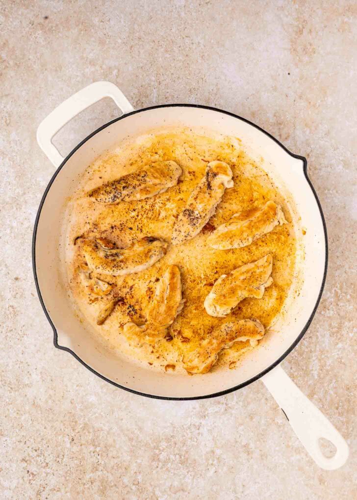 Cooked seasoned chicken strips in a white skillet on a light-colored countertop. The chicken is browned and surrounded by juices and spices.
