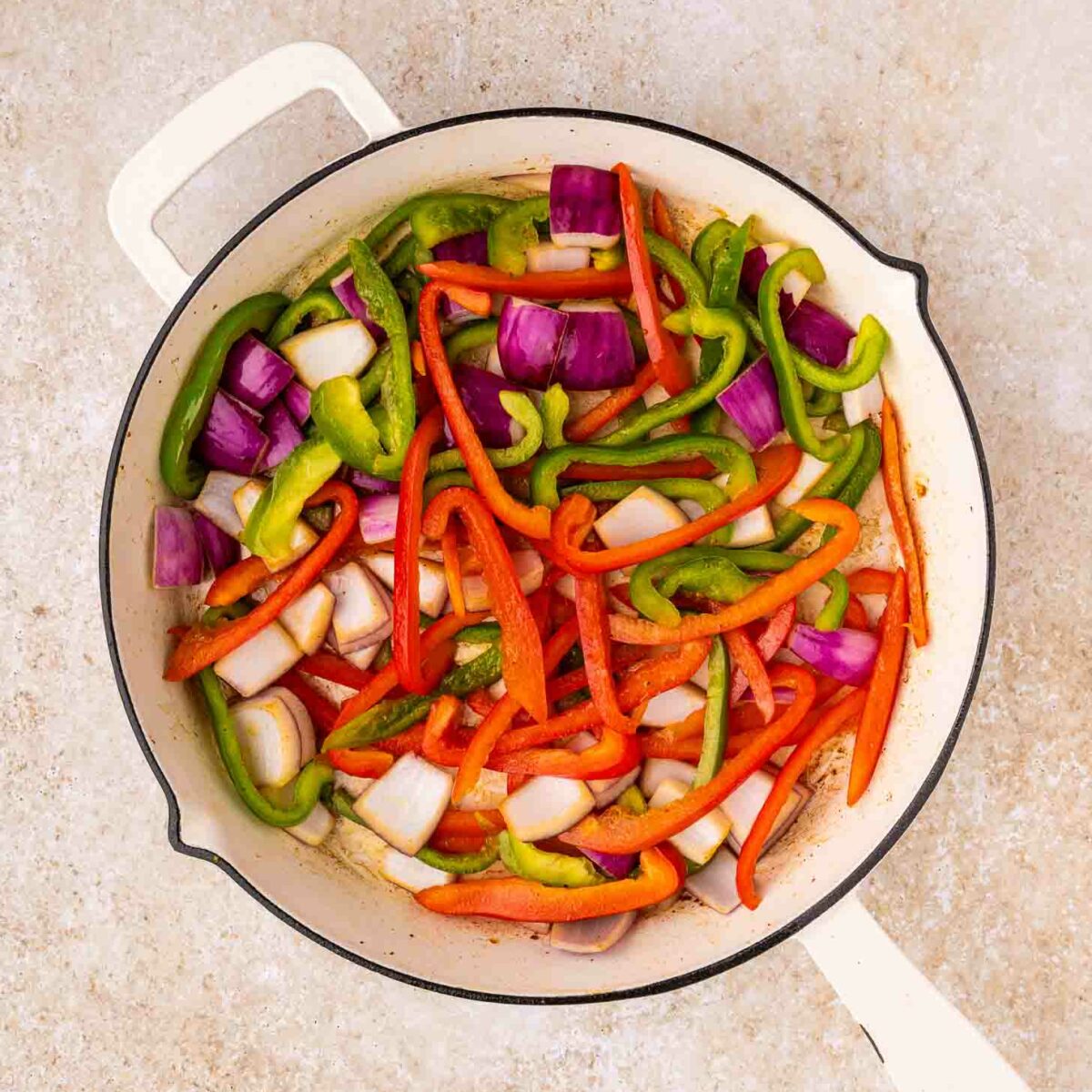 A white skillet filled with sliced red and green bell peppers and chunks of red onion, arranged on a light-colored countertop.