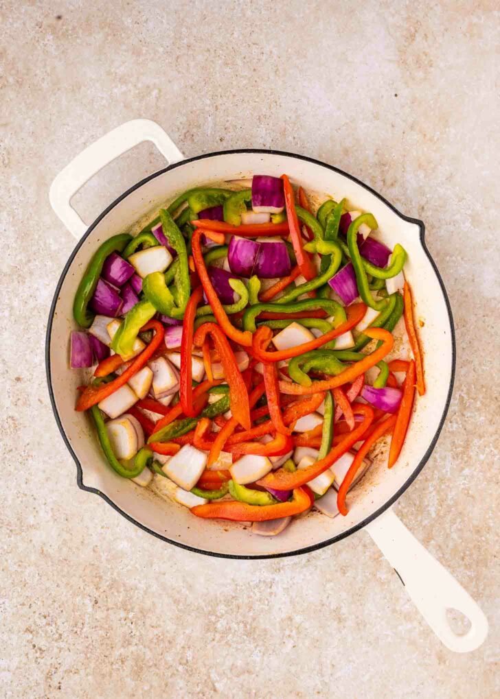 A white skillet filled with sliced red and green bell peppers and chunks of red onion, arranged on a light-colored countertop.