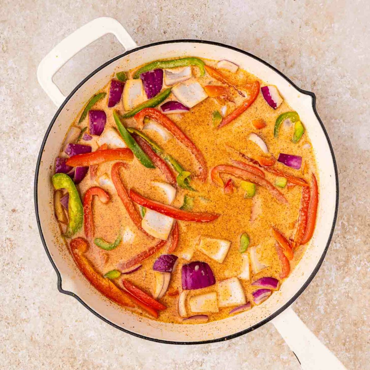 A white skillet filled with a creamy orange sauce, sliced red onions, red bell peppers, and green bell peppers, sitting on a light stone countertop.