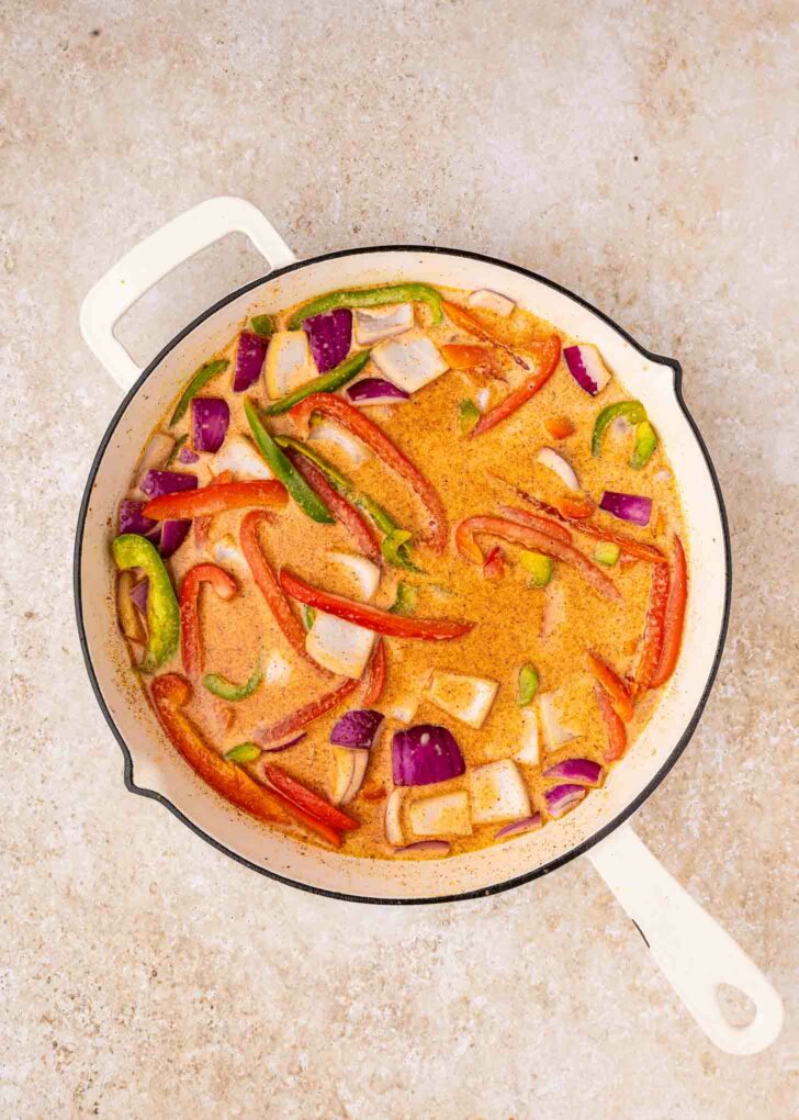 A white skillet filled with a creamy orange sauce, sliced red onions, red bell peppers, and green bell peppers, sitting on a light stone countertop.