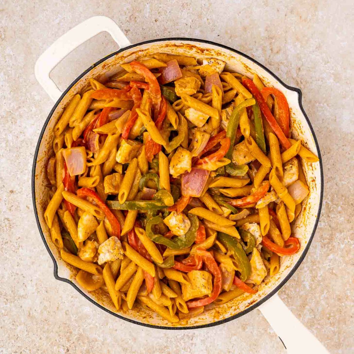 A white skillet filled with cooked penne pasta, diced chicken, red and green bell peppers, and red onions in a seasoned sauce, set on a light-colored countertop.