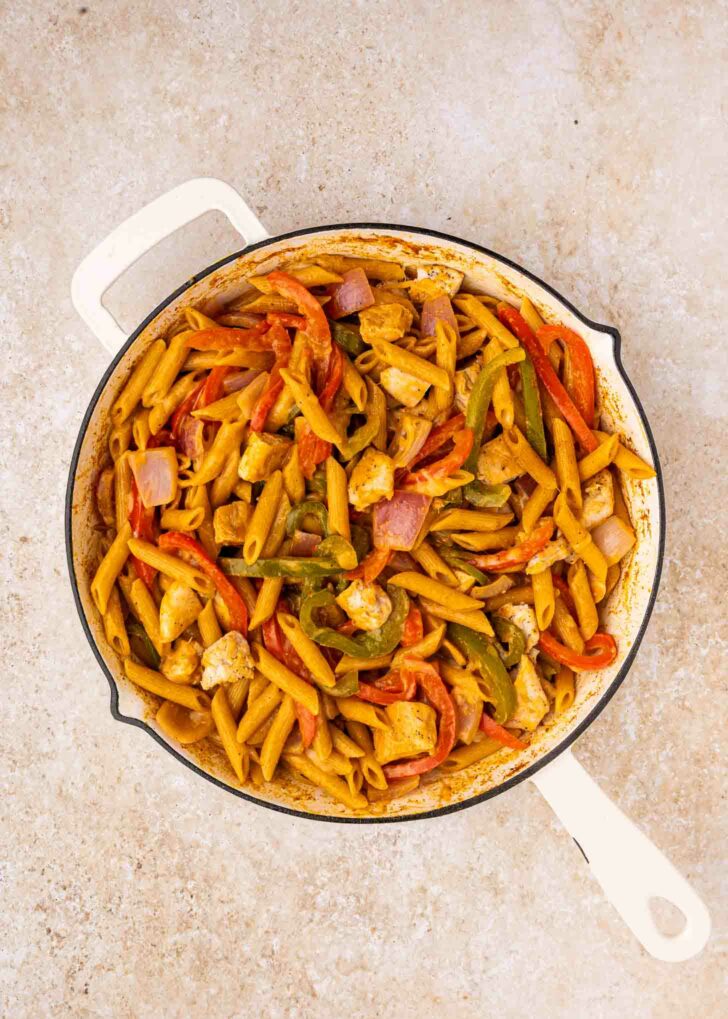 A white skillet filled with cooked penne pasta, diced chicken, red and green bell peppers, and red onions in a seasoned sauce, set on a light-colored countertop.