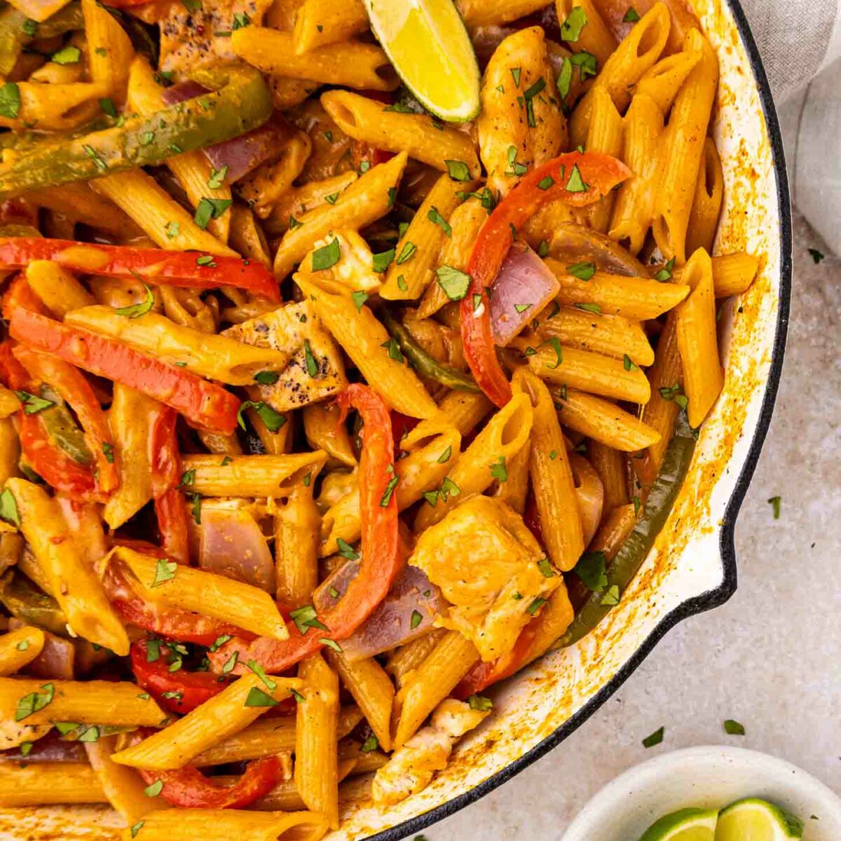 A skillet filled with penne pasta, cooked with sliced red and green bell peppers, onions, and pieces of chicken, garnished with fresh herbs and lime wedges. A small bowl with extra lime wedges is nearby.