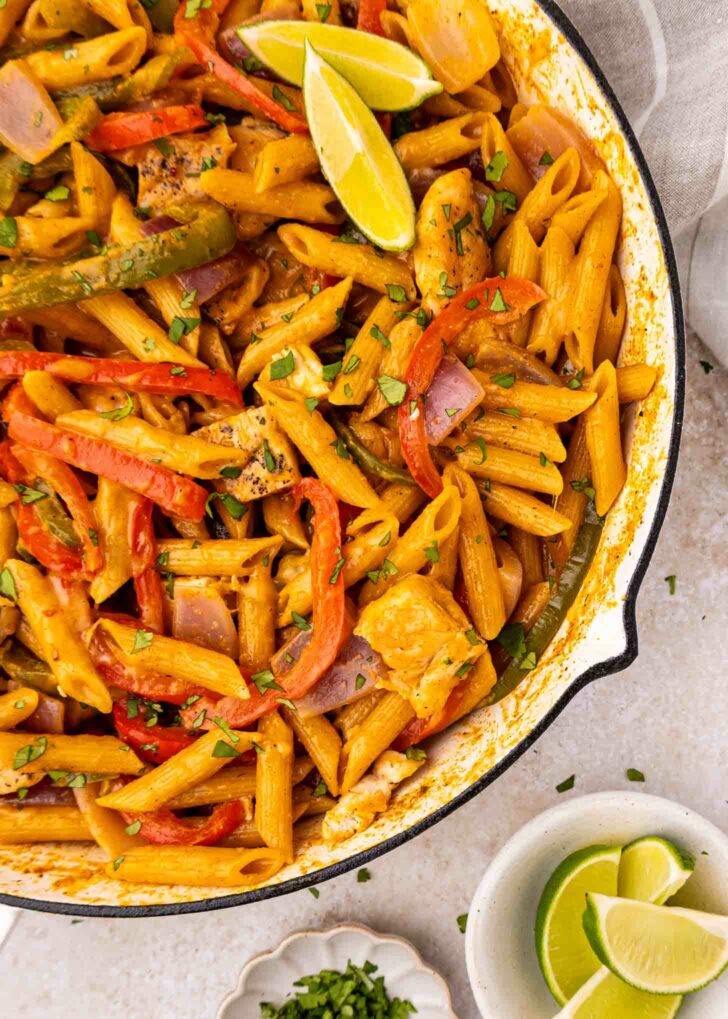 A skillet filled with penne pasta, cooked with sliced red and green bell peppers, onions, and pieces of chicken, garnished with fresh herbs and lime wedges. A small bowl with extra lime wedges is nearby.