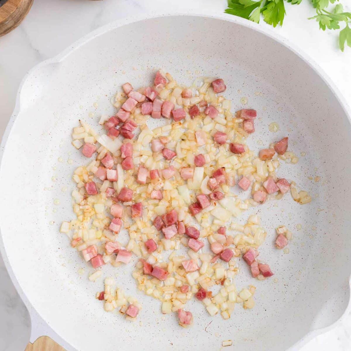 A white skillet with diced onions and pancetta sautéing, on a marble countertop. Nearby are parsley sprigs, a wooden pepper grinder, and a small bowl of black peppercorns.