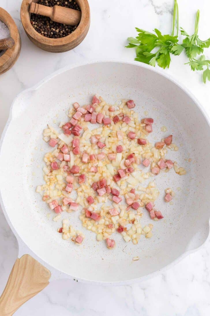 A white skillet with diced onions and pancetta sautéing, on a marble countertop. Nearby are parsley sprigs, a wooden pepper grinder, and a small bowl of black peppercorns.