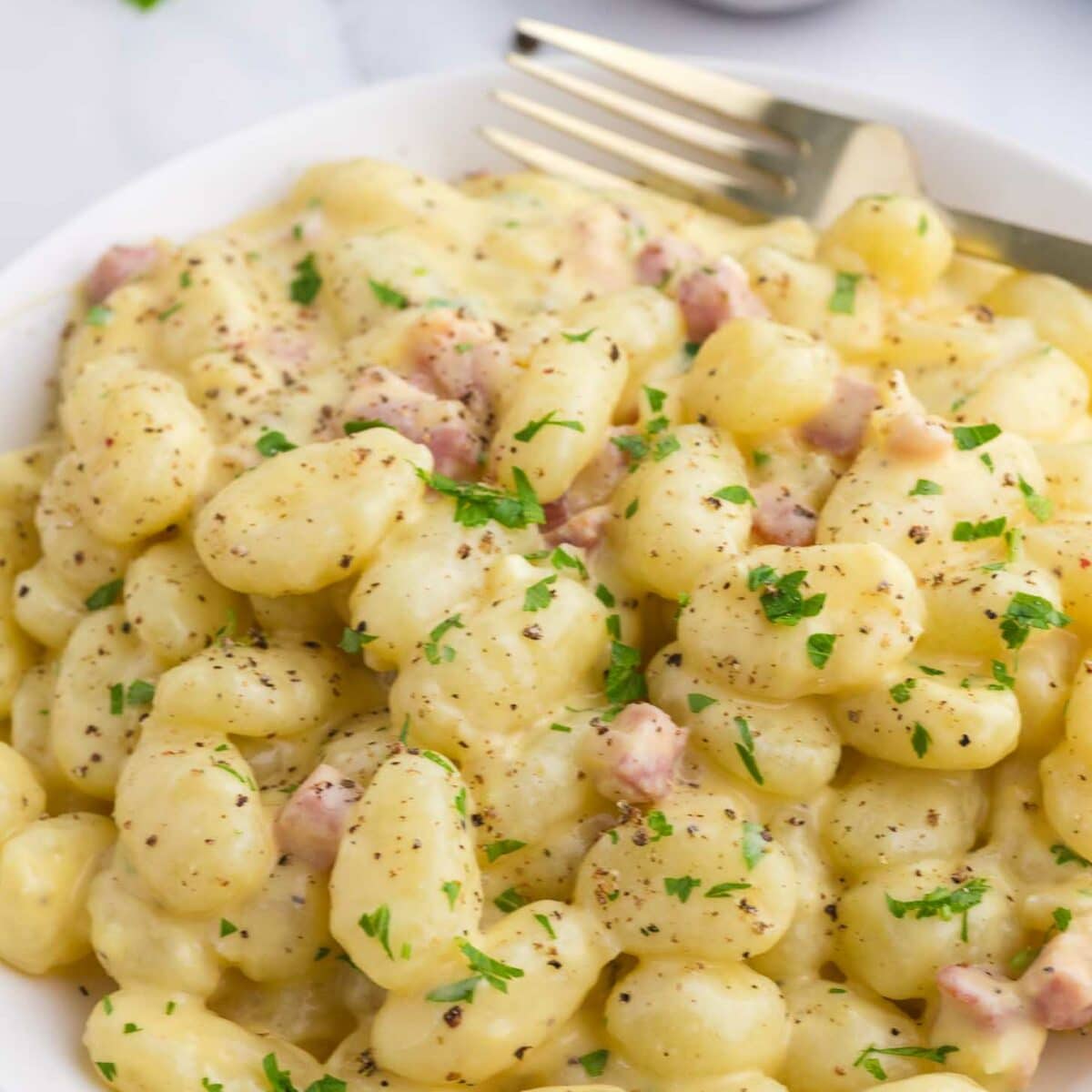 A white plate of creamy gnocchi with diced pancetta, garnished with chopped parsley and cracked black pepper. A fork rests on the side, and a small bowl of grated cheese is visible in the background.