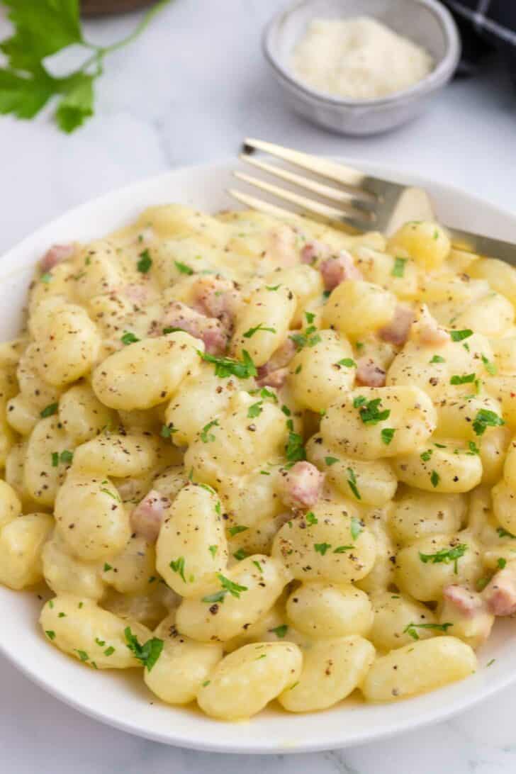 A white plate of creamy gnocchi with diced pancetta, garnished with chopped parsley and cracked black pepper. A fork rests on the side, and a small bowl of grated cheese is visible in the background.