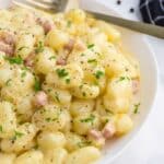 A white bowl filled with creamy gnocchi, garnished with chopped parsley and black pepper, sits on a marble surface next to a gold fork and a black checkered napkin.