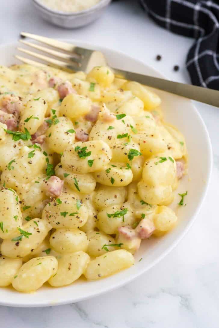 A white bowl filled with creamy gnocchi, garnished with chopped parsley and black pepper, sits on a marble surface next to a gold fork and a black checkered napkin.