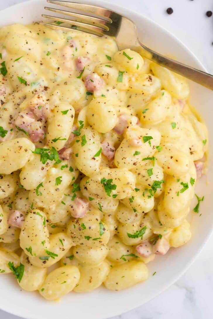 A bowl of creamy gnocchi topped with diced pancetta, black pepper, and chopped parsley, with a fork resting on the side.