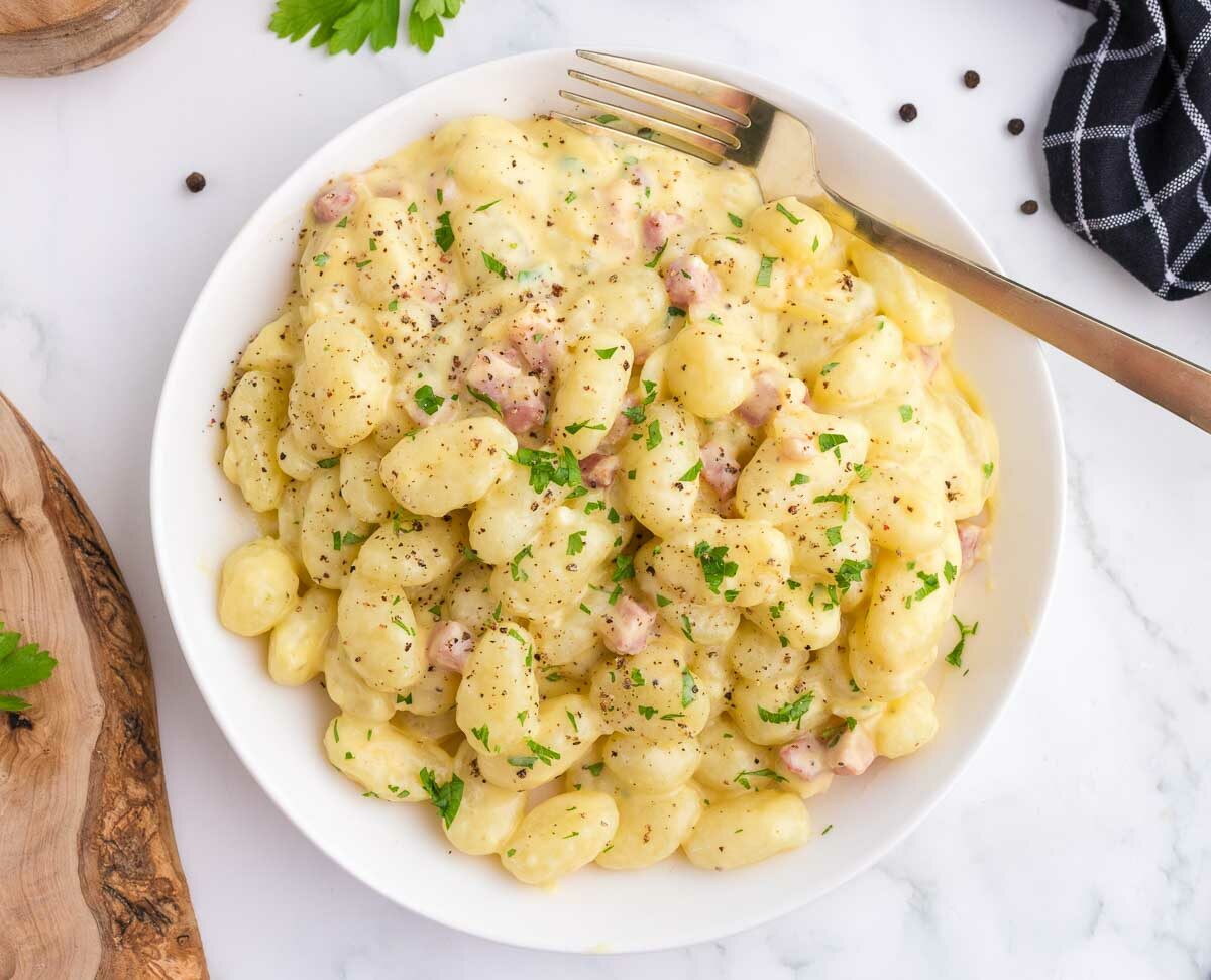 A white plate of creamy gnocchi with pieces of pancetta, topped with chopped parsley and black pepper. A fork rests on the plate, and a black checkered napkin is nearby on a white surface.