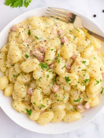 A white plate of creamy gnocchi with pieces of pancetta, topped with chopped parsley and black pepper. A fork rests on the plate, and a black checkered napkin is nearby on a white surface.