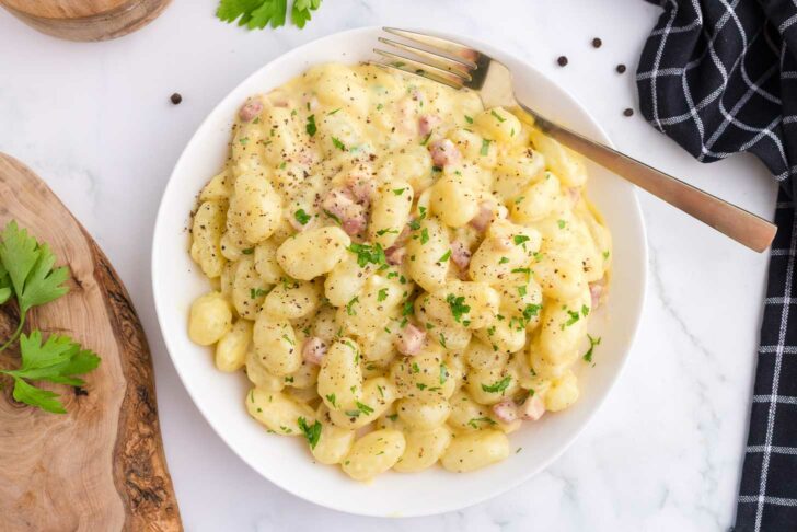 A white plate of creamy gnocchi with pieces of pancetta, topped with chopped parsley and black pepper. A fork rests on the plate, and a black checkered napkin is nearby on a white surface.