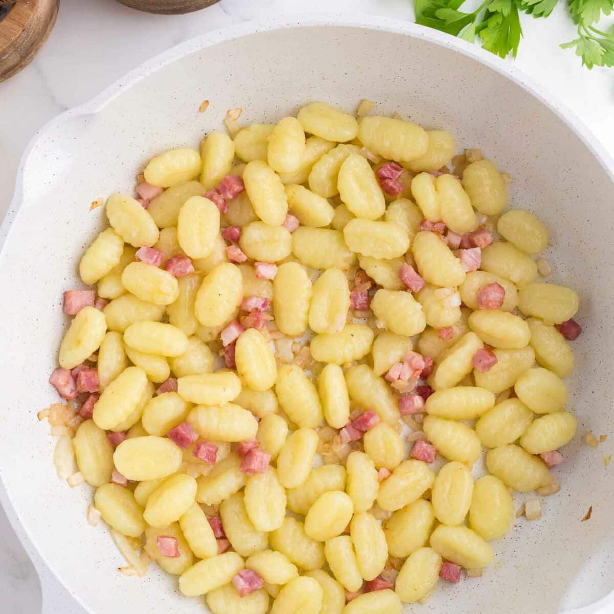 A white skillet filled with cooked gnocchi and small diced pieces of pancetta. Nearby are wooden bowls with black peppercorns and parsley leaves on a marble countertop.
