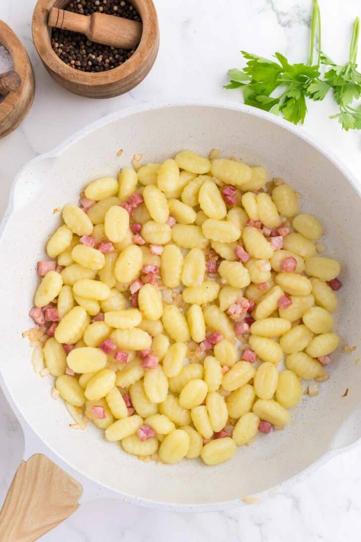 A white skillet filled with cooked gnocchi and small diced pieces of pancetta. Nearby are wooden bowls with black peppercorns and parsley leaves on a marble countertop.