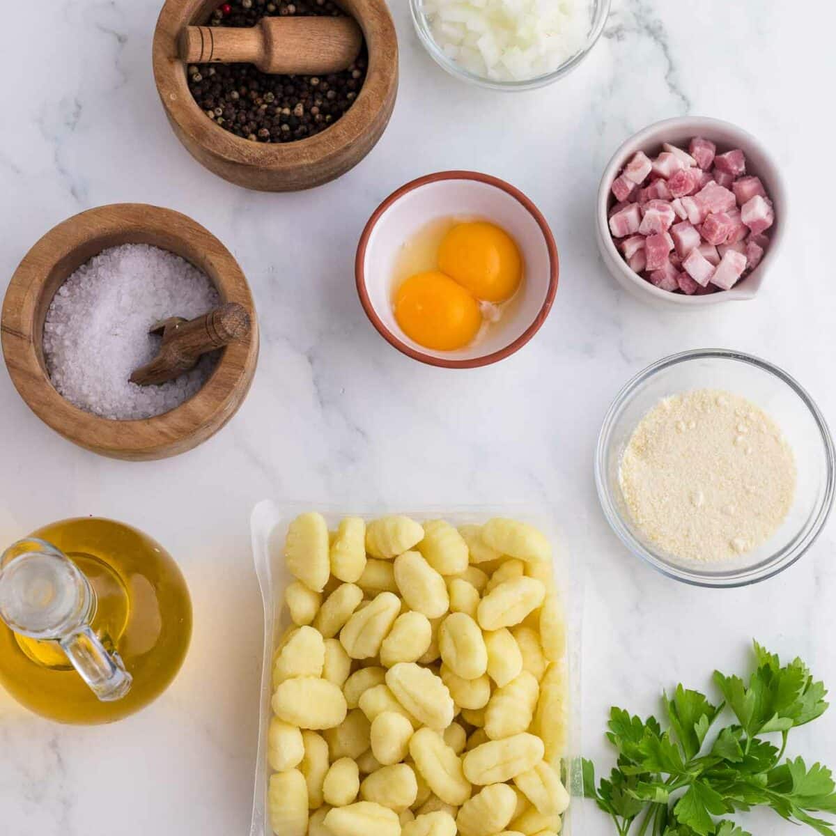 Ingredients on a white marble surface: gnocchi, parsley, olive oil, salt, peppercorns with mortar, chopped onion, two eggs, diced pancetta, and grated cheese in small bowls.