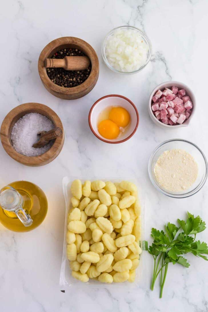 Ingredients on a white marble surface: gnocchi, parsley, olive oil, salt, peppercorns with mortar, chopped onion, two eggs, diced pancetta, and grated cheese in small bowls.