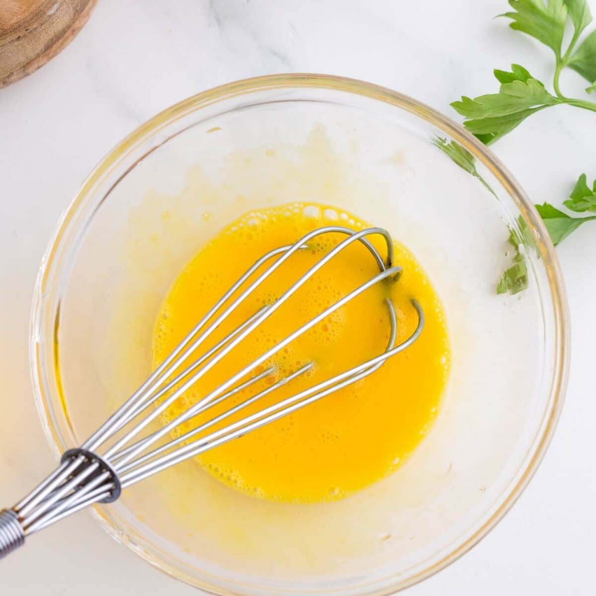 A metal whisk in a glass bowl containing beaten eggs sits on a white surface, with a sprig of parsley and a wooden bowl nearby.