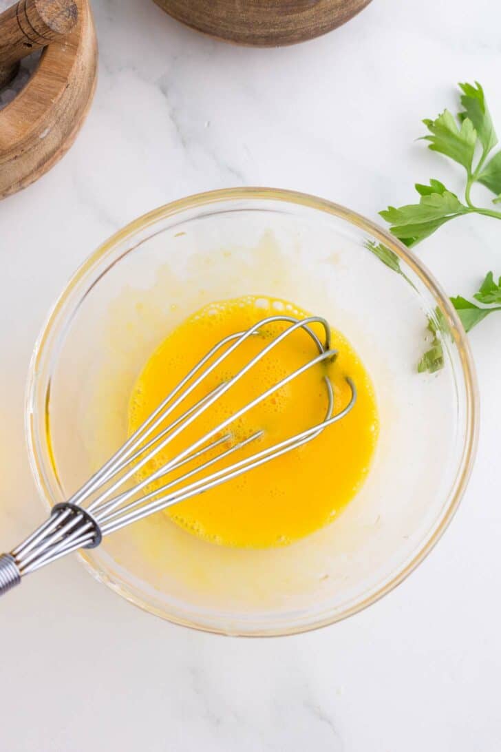 A metal whisk in a glass bowl containing beaten eggs sits on a white surface, with a sprig of parsley and a wooden bowl nearby.