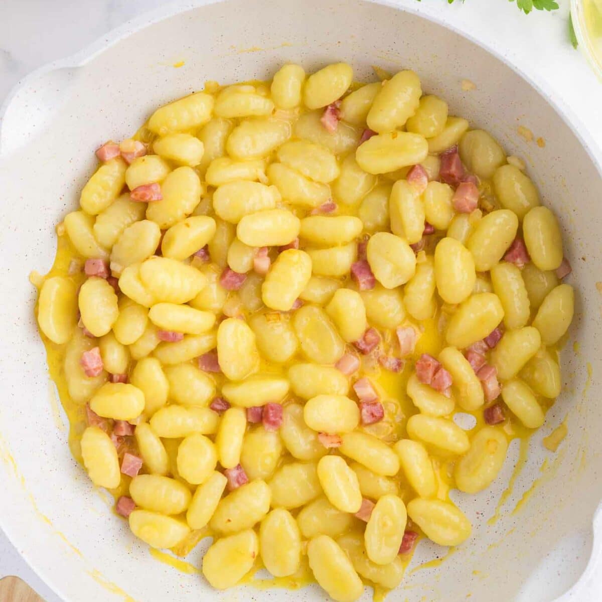 A white skillet filled with gnocchi mixed with small cubes of pancetta in a creamy yellow sauce, with parsley and a pepper grinder nearby on a white surface.