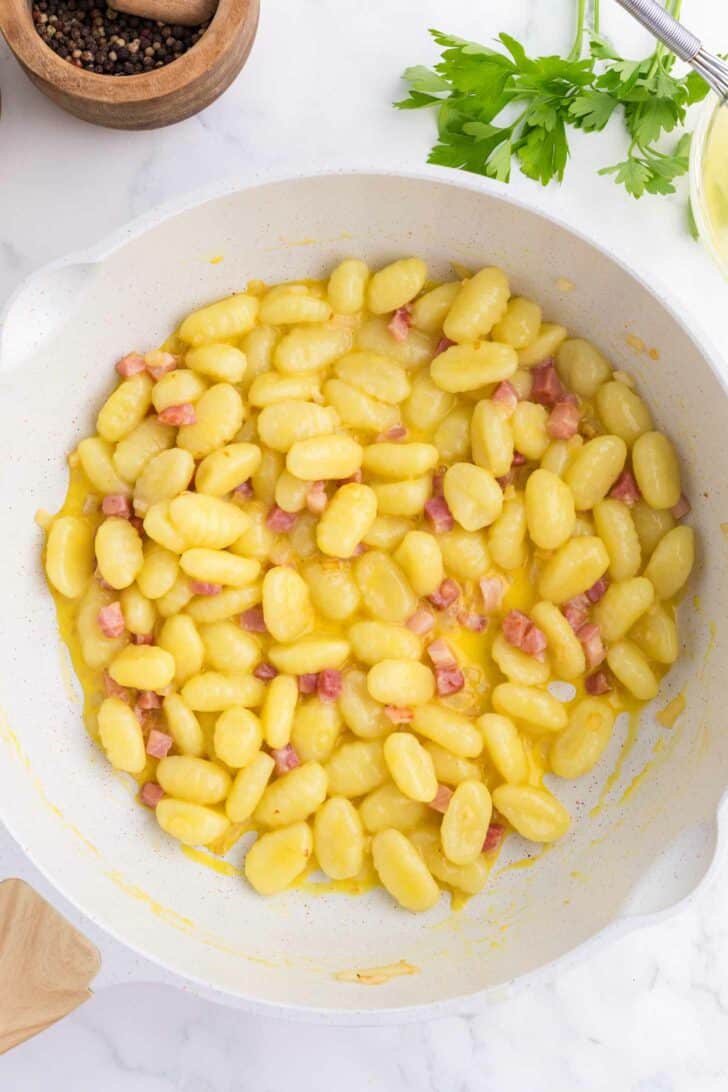 A white skillet filled with gnocchi mixed with small cubes of pancetta in a creamy yellow sauce, with parsley and a pepper grinder nearby on a white surface.