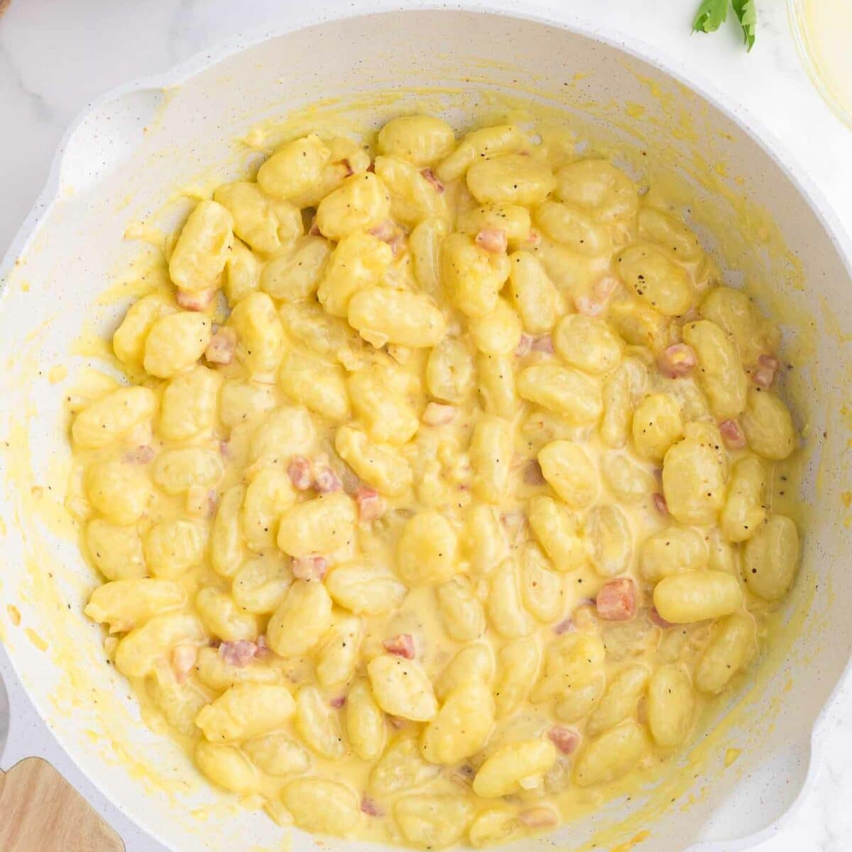 A white skillet filled with creamy gnocchi mixed with small pieces of pancetta, garnished with black pepper. Fresh parsley and kitchen items are visible in the background.