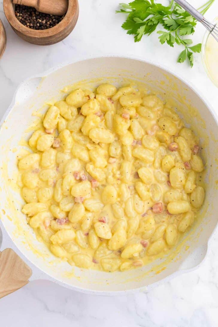 A white skillet filled with creamy gnocchi mixed with small pieces of pancetta, garnished with black pepper. Fresh parsley and kitchen items are visible in the background.