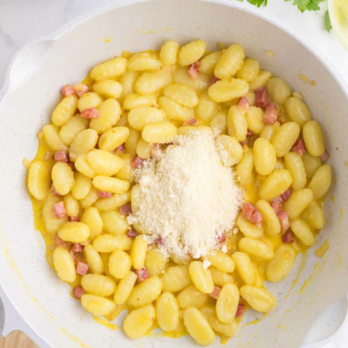 A white skillet filled with cooked gnocchi, small pieces of pancetta, and a mound of grated parmesan cheese on top, with parsley, pepper, and a whisk visible in the background.