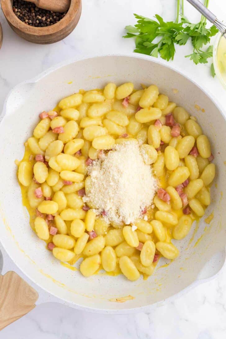 A white skillet filled with cooked gnocchi, small pieces of pancetta, and a mound of grated parmesan cheese on top, with parsley, pepper, and a whisk visible in the background.