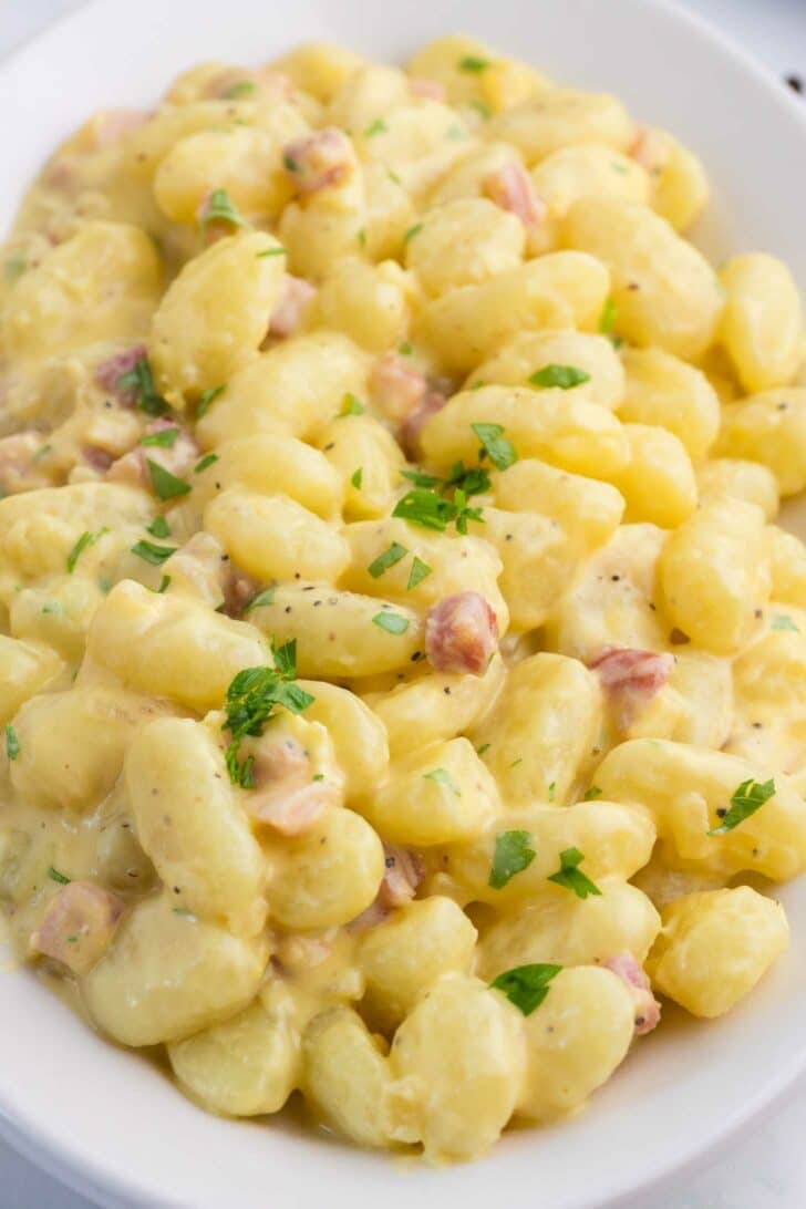 A white plate filled with creamy gnocchi in a cream sauce, garnished with chopped parsley and small pieces of pancetta.