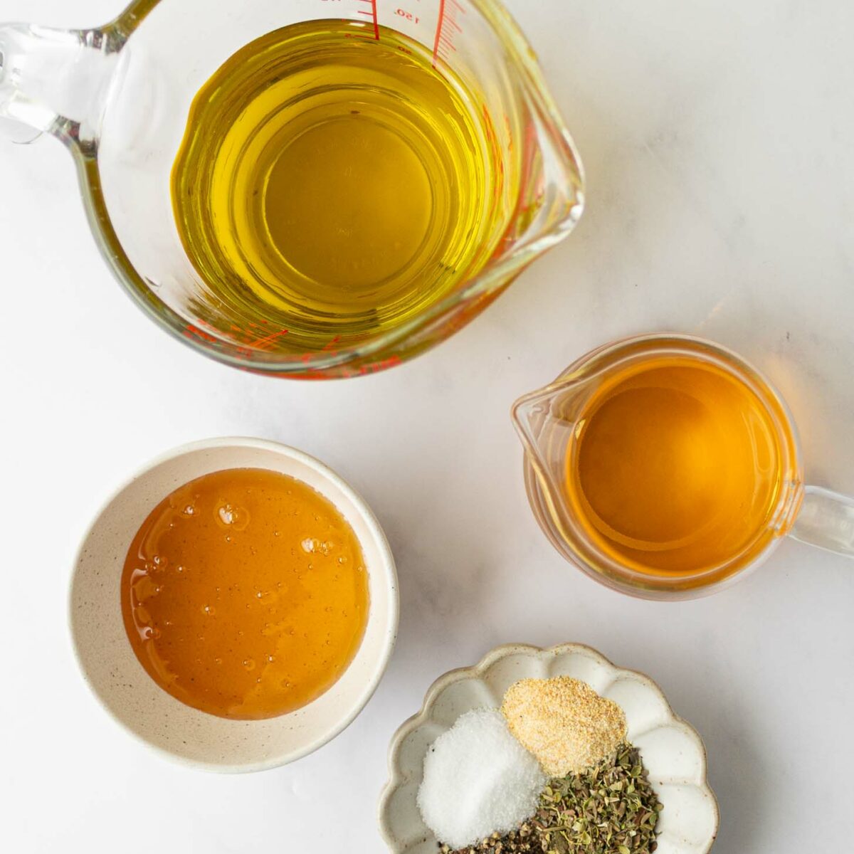 Four containers on a white surface: a measuring cup of olive oil, a small pitcher of white balsamic vinegar, a bowl of honey, and a dish with dried herbs, salt, pepper, and garlic powder.