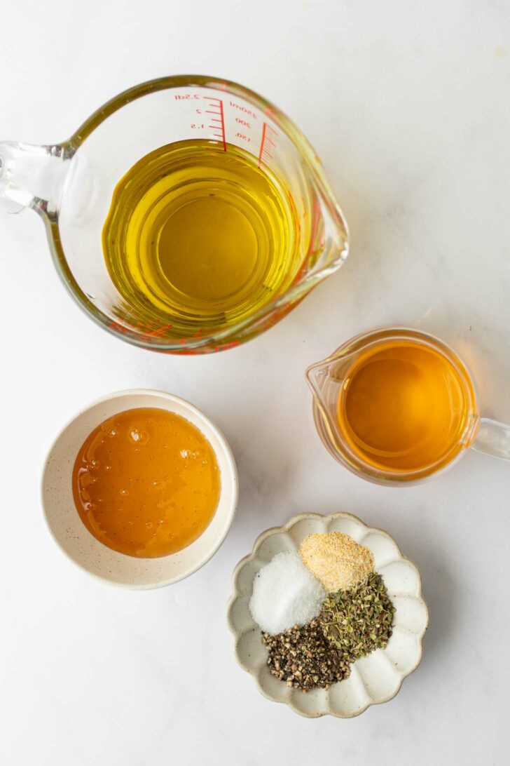 Four containers on a white surface: a measuring cup of olive oil, a small pitcher of white balsamic vinegar, a bowl of honey, and a dish with dried herbs, salt, pepper, and garlic powder.