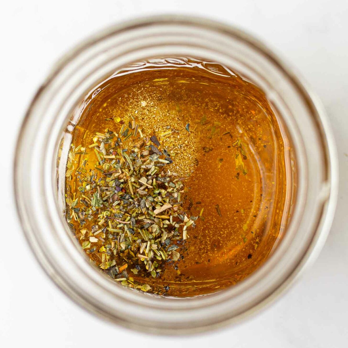 A glass jar filled with golden liquid with dried herbs sprinkled on top, viewed from above on a white surface.