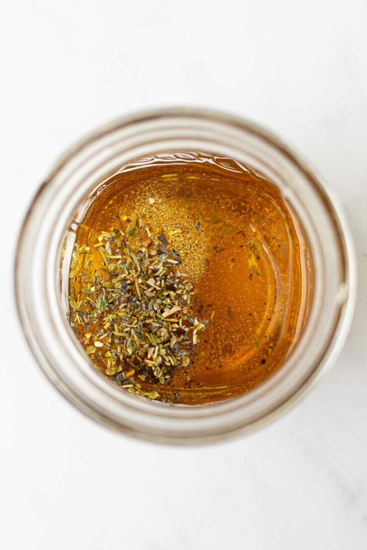 A glass jar filled with golden liquid with dried herbs sprinkled on top, viewed from above on a white surface.