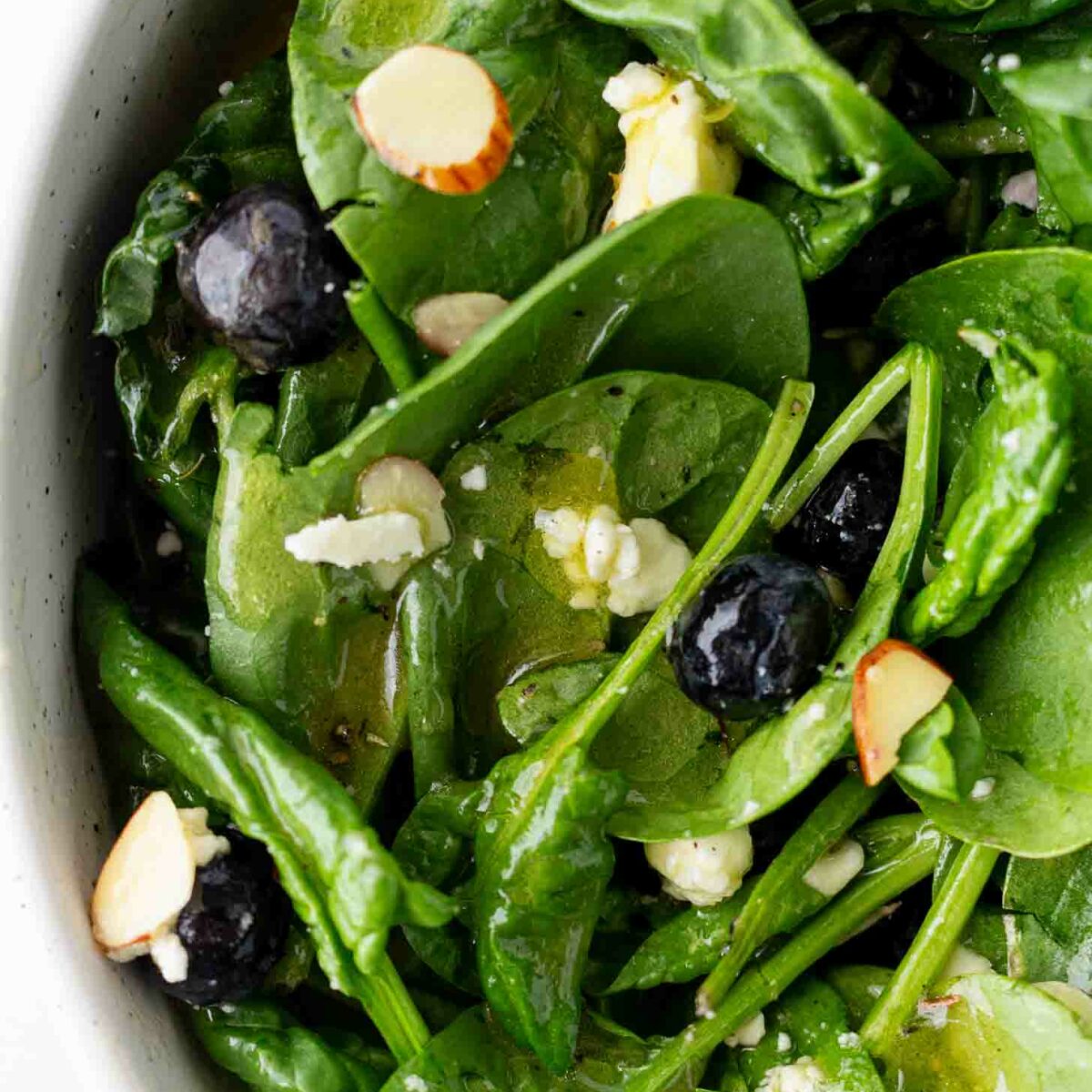 Fresh spinach salad with blueberries, sliced almonds, and crumbled cheese, lightly dressed, shown in a close-up view inside a white bowl.