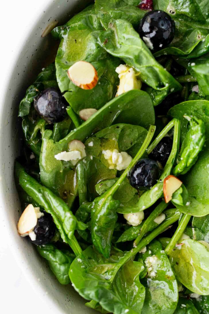 Fresh spinach salad with blueberries, sliced almonds, and crumbled cheese, lightly dressed, shown in a close-up view inside a white bowl.