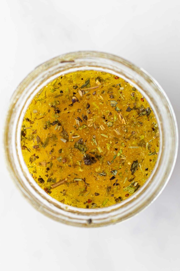 A top-down view of a jar filled with dressing mixed with herbs and spices, set against a light background.