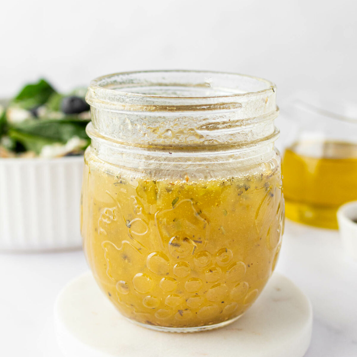 A glass jar filled with salad dressing sits on a round white coaster, with a salad bowl and a small glass of oil blurred in the background.
