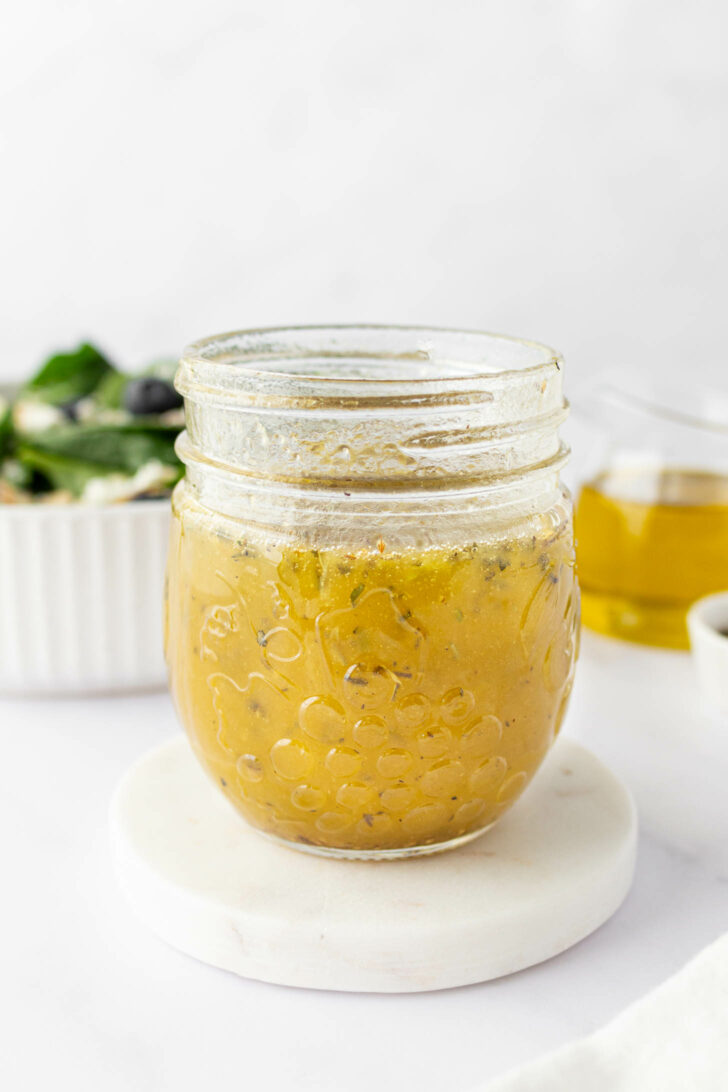 A glass jar filled with salad dressing sits on a round white coaster, with a salad bowl and a small glass of oil blurred in the background.