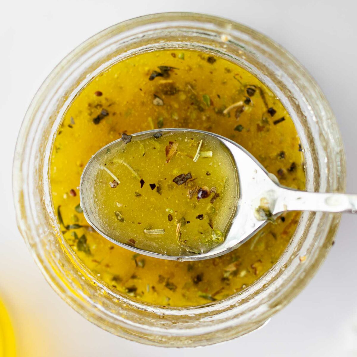 A close-up, top-down view of a jar filled with dressing, with a spoon resting on top holding some of the yellow, herb-speckled liquid.