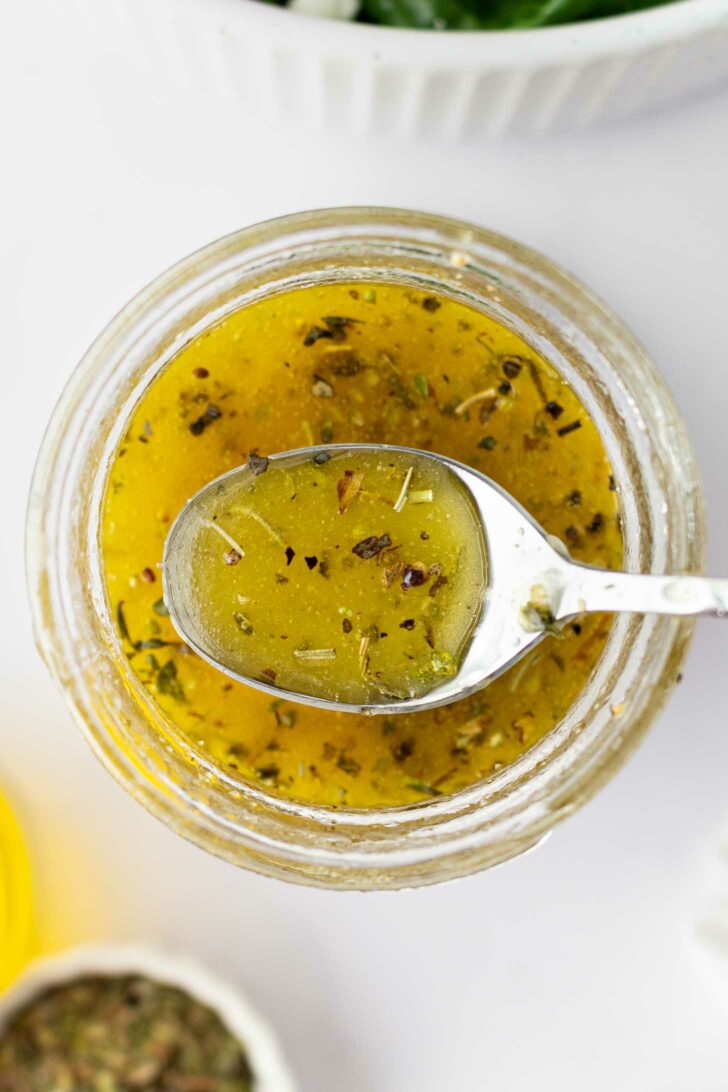 A close-up, top-down view of a jar filled with dressing, with a spoon resting on top holding some of the yellow, herb-speckled liquid.