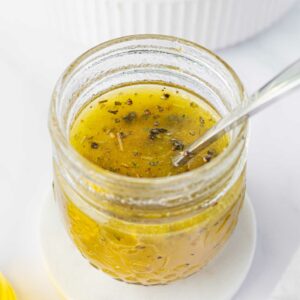 A glass jar filled with yellowish homemade salad dressing with herbs, a spoon inside, placed on a white coaster. In the background, there is a green salad in a white bowl.