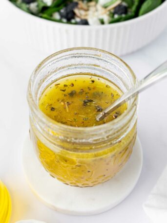 A glass jar filled with yellowish homemade salad dressing with herbs, a spoon inside, placed on a white coaster. In the background, there is a green salad in a white bowl.