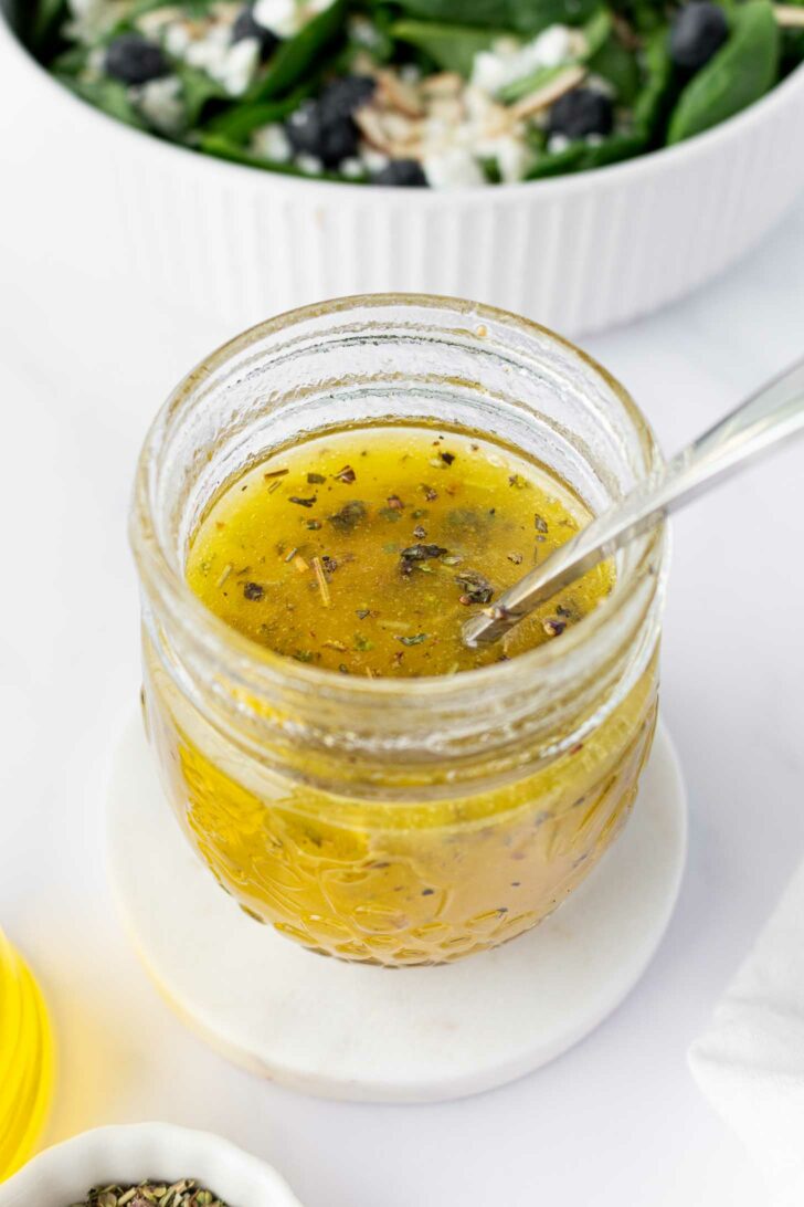 A glass jar filled with yellowish homemade salad dressing with herbs, a spoon inside, placed on a white coaster. In the background, there is a green salad in a white bowl.