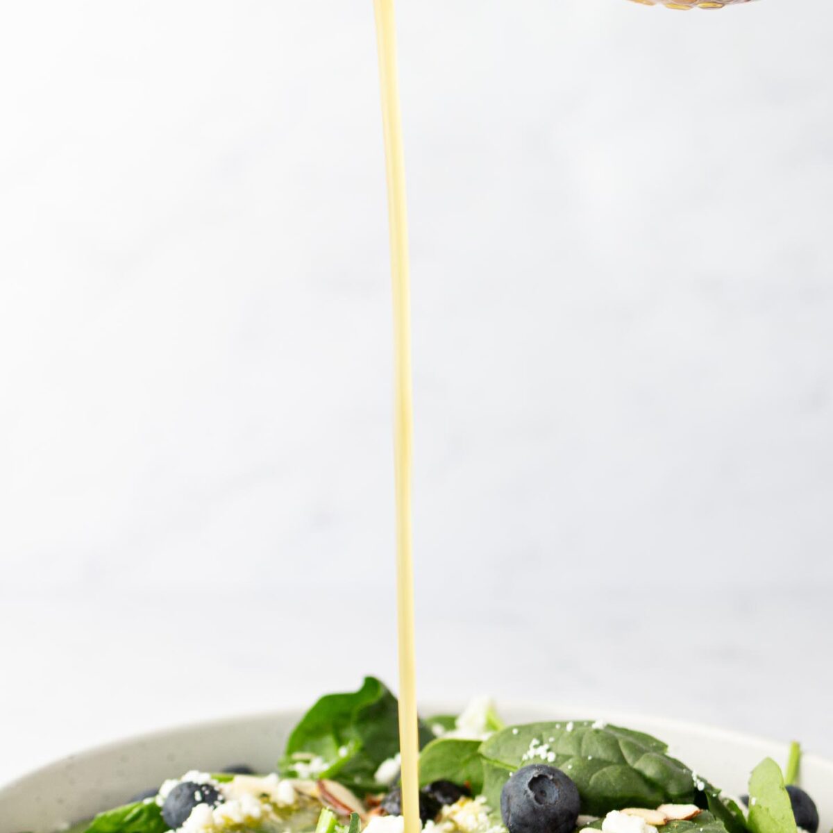 A hand pours salad dressing from a jar onto a bowl of fresh spinach salad topped with blueberries, crumbled cheese, and almond slices.