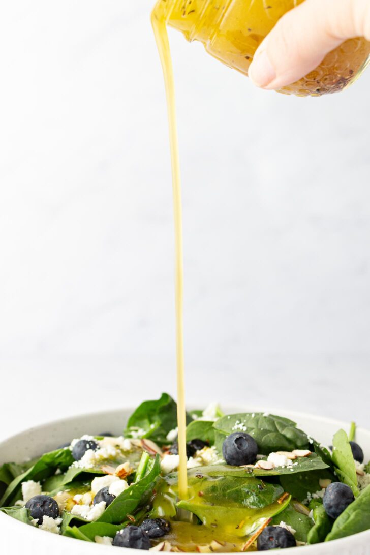 A hand pours salad dressing from a jar onto a bowl of fresh spinach salad topped with blueberries, crumbled cheese, and almond slices.