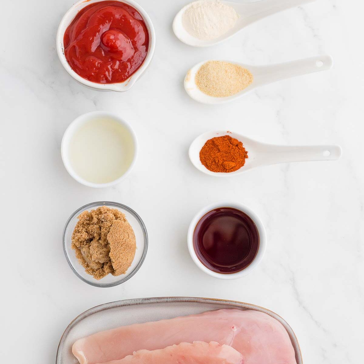 Flat lay of raw chicken breasts, ketchup, vinegar, brown sugar, onion powder, garlic powder, paprika, and spices in bowls and spoons on a white marble surface.